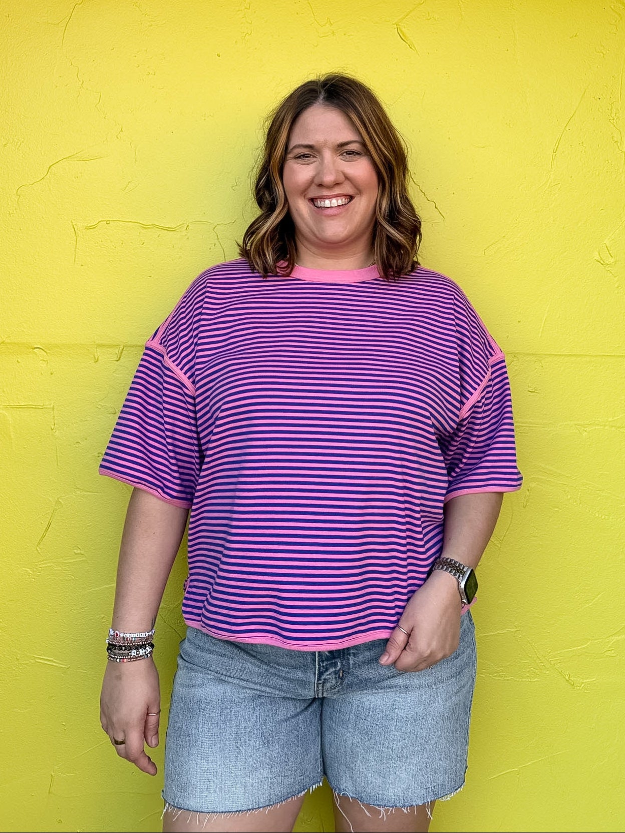 Person wearing a purple striped shirt and denim shorts standing against a bright yellow wall.