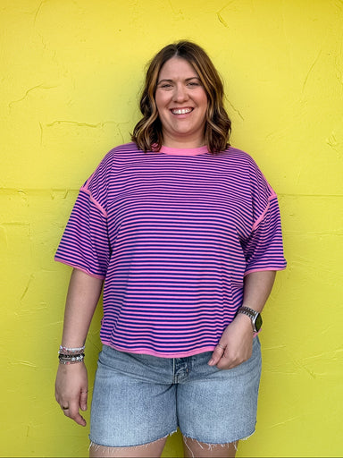 Person wearing a purple striped shirt and denim shorts standing against a bright yellow wall.