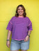 Person wearing a purple striped shirt and denim shorts standing against a bright yellow wall.